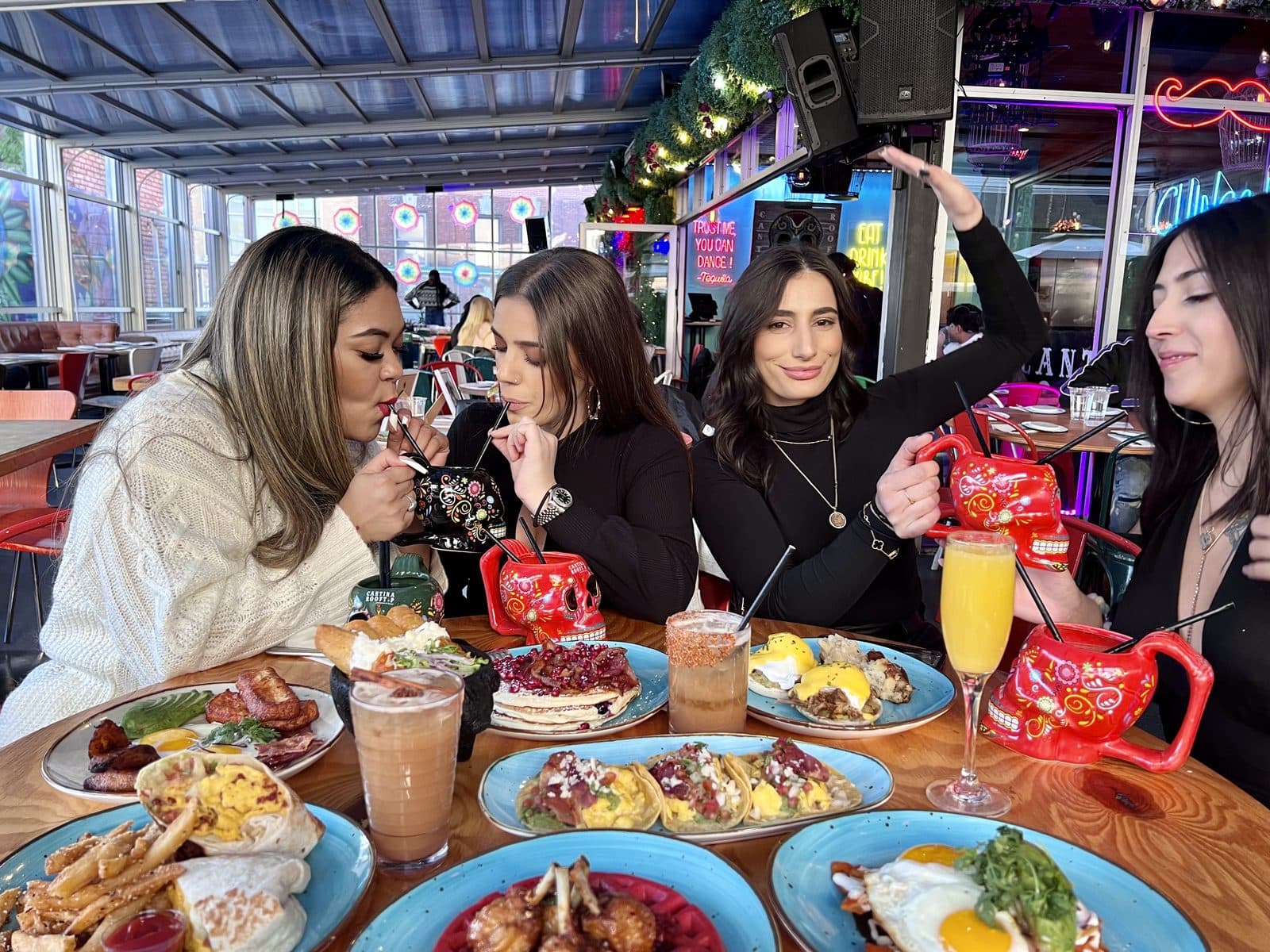 Four friends sharing brunch plates and red skull mugs at a table under the neon lights