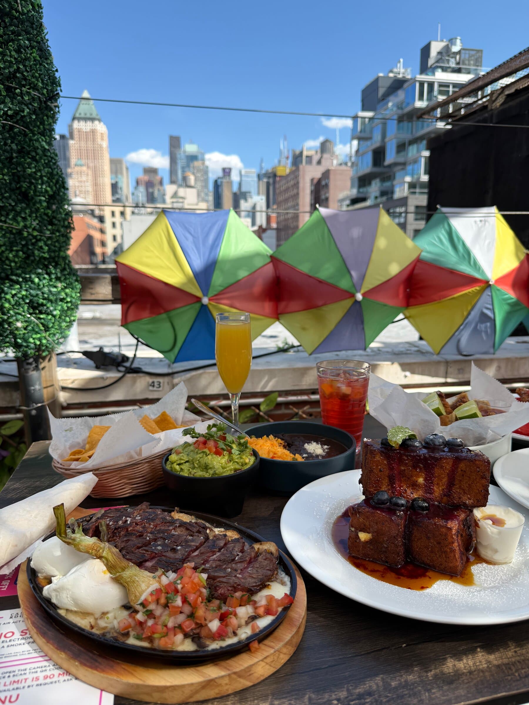 Brunch plates and drinks on a rooftop table, with rainbow umbrellas and the city behind