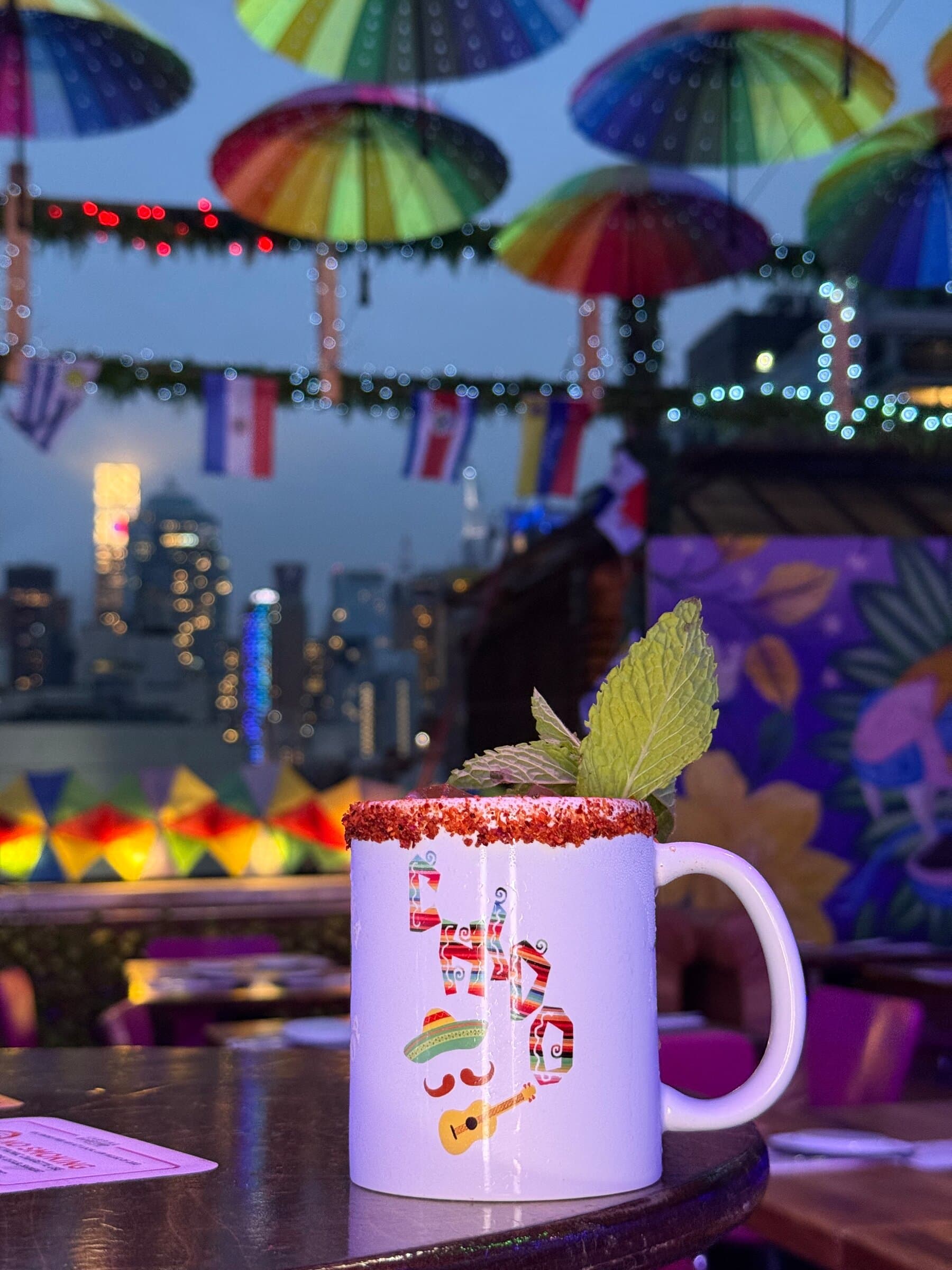 A chili-rimmed cocktail with mint on a rooftop table at dusk, under rainbow umbrellas and string lights