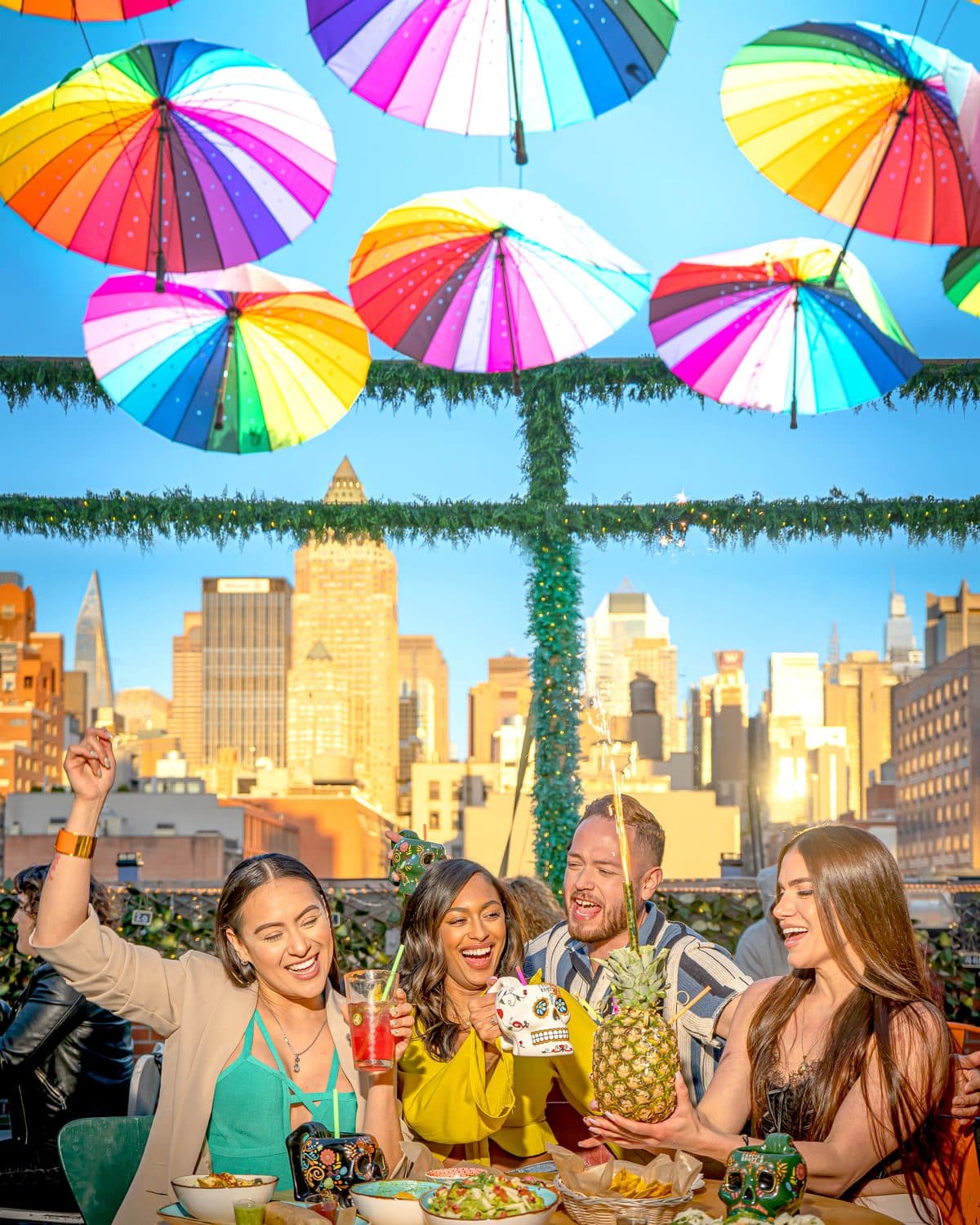 Friends toasting with skull mugs and a piña loca under the rainbow umbrellas, Midtown skyline behind