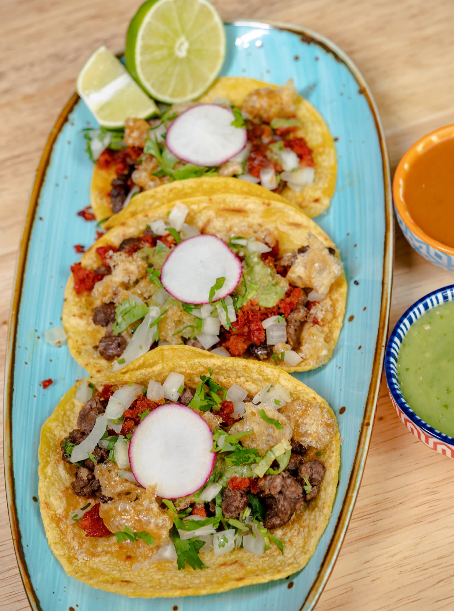 Three tacos on corn tortillas with onion, cilantro and radish on a blue plate, with lime and two salsas